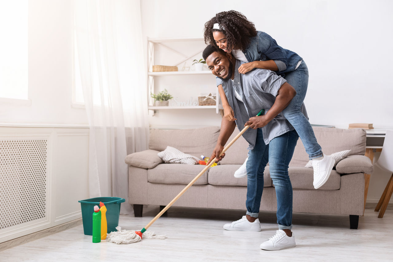 A man gives a piggyback ride to a smiling woman while he holds a mop in a clean room.