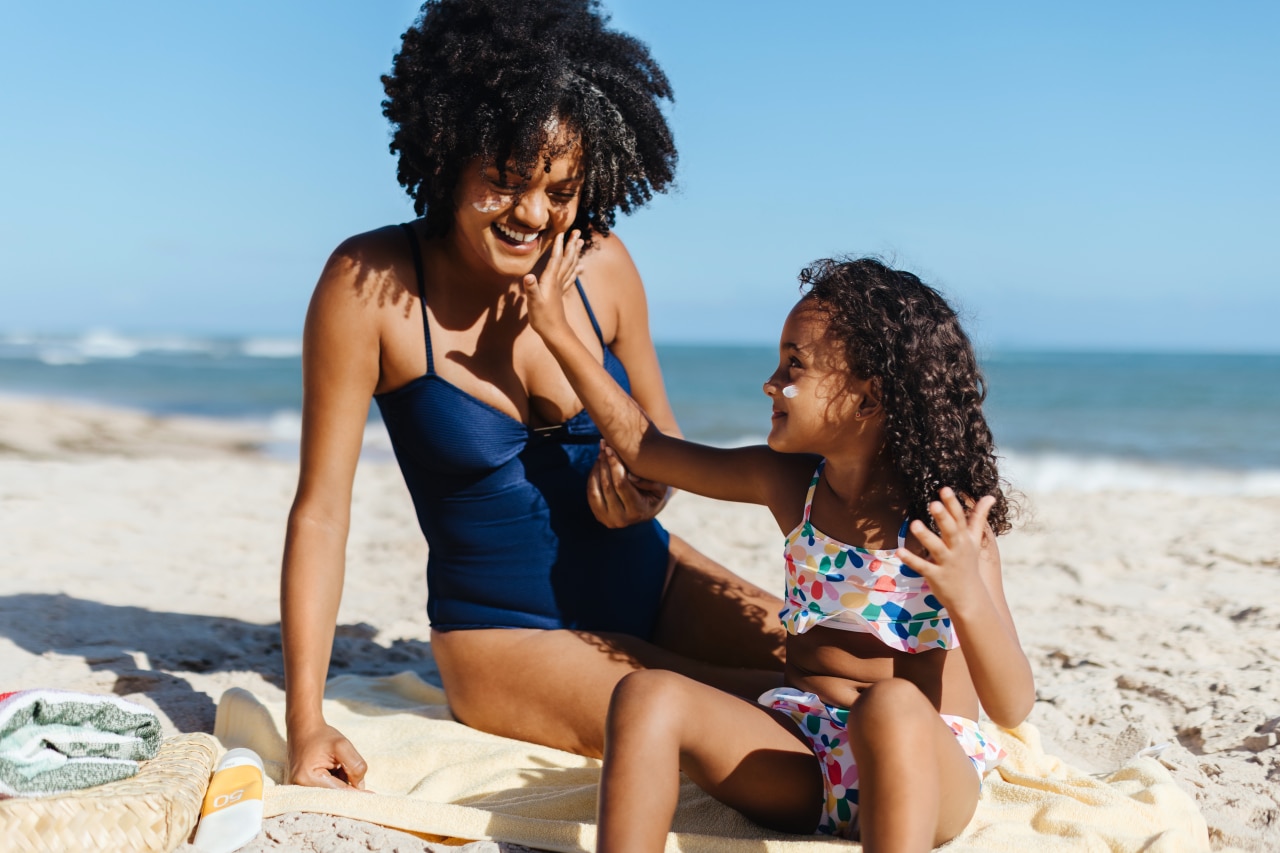 Child playfully applying sunscreen on mother's face on beach for sun protection.