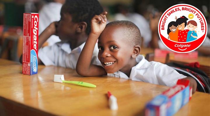 Smiling child in a classroom setting with Colgate toothpaste and toothbrush on the desk, park of Colgate's Bright Smiles, Bright Futures oral health education program.