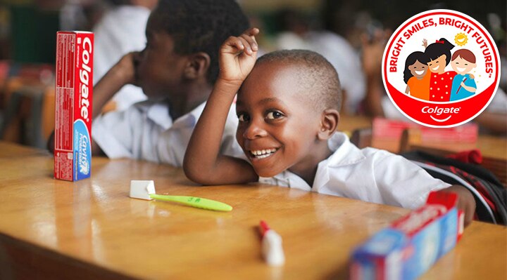 Smiling child in a classroom setting with Colgate toothpaste and toothbrush on the desk, park of Colgate's Bright Smiles, Bright Futures oral health education program.