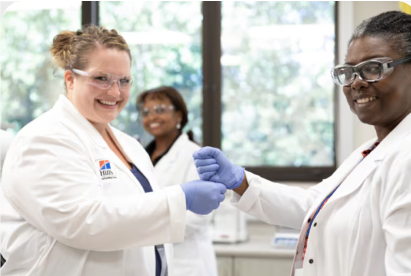 Two scientists in lab coats and gloves smile and shake hands in a laboratory setting.