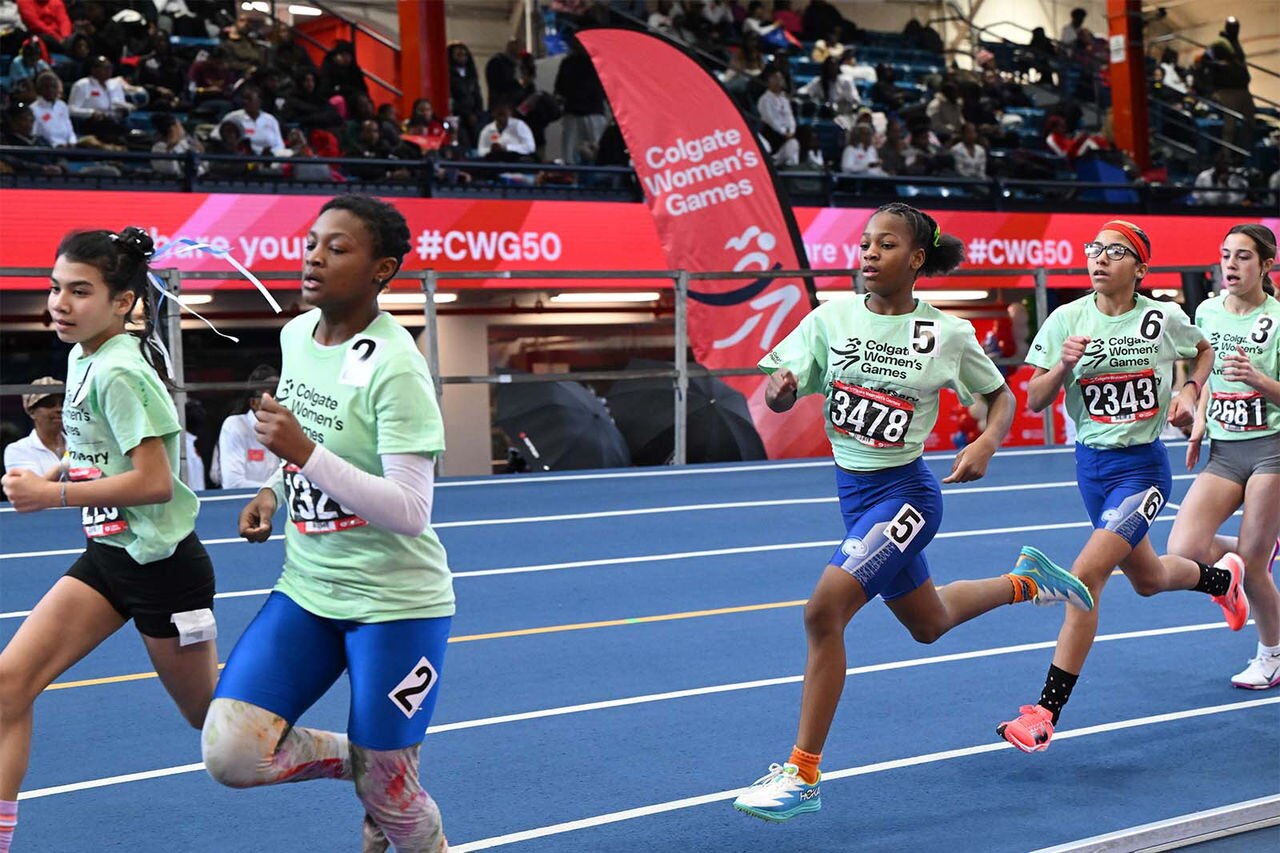 A group of young female athletes sprint together along a blue indoor track during a track and field race.﻿