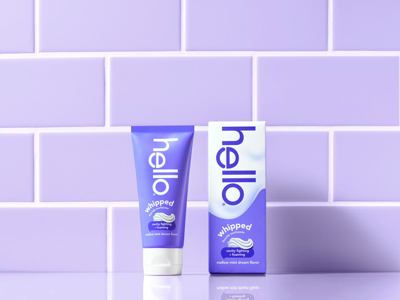 Purple and white hello Whipped toothpaste tube and box in front of purple tiled wall.