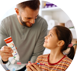 A man applying toothpaste to a girl's toothbrush at a sink, with plants in the background.